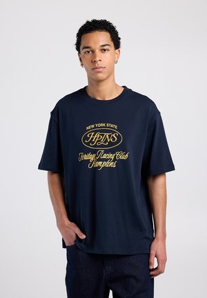 T-shirt en coton bleu marine à manches courtes, avec un motif jaune indiquant "New York State HLNS Heritage Racing Club Hamptons."