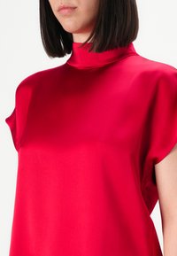 Femme portant un haut sans manches en satin rouge vif à col haut, avec des cheveux bruns foncés droits coupés au niveau du menton, sur fond blanc.