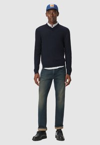 Suéter de cuello en v navy, camisa de botones azul claro, jeans oscuros y zapatos negros con cordones. El modelo lleva una gorra azul con un logo.