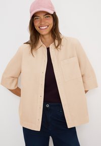 Mujer sonriente que lleva una gorra rosa, una camisa beige con botones y bolsillo, un top oscuro y jeans oscuros frente a un fondo blanco liso.