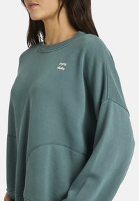 Sudadera en color verde azulado, fabricada con tela suave. Presenta un cuello acanalado, hombros caídos y un pequeño logo en el pecho.