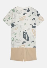 Kreminės spalvos trumpomis rankovėmis marškinėliai su daugiakulūnėmis Nike logotipo spaudėmis; derinami su šviesiai rudais šortais, pagamintais iš minkštos medžiagos, patogaus kirpimo.