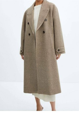 Personne portant un long manteau en laine beige croisé sur une robe en maille blanche, debout devant un fond blanc uni.