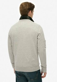Hombre de espaldas, vistiendo un sudadero gris claro con cuello a cuadros verde oscuro y jeans azules, frente a un fondo blanco liso.