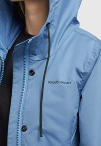 Ljusblå jacka med huva, med dragkedja framtill, tryckknappsstängning och svart dragsko. Logotyp tryckt på bröstet. Mjuk textur.