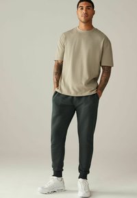 Helle beige kurzärmelige T-Shirt, lockere Passform, kombiniert mit dunkelgrünen Jogginghosen mit gerippten Bündchen, ergänzt durch weiße Sportschuhe.