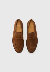 Mocassins en daim marron avec un bout arrondi, des coutures apparentes et une lanière décorative sur le dessus. Semelle intérieure en cuir, semelle fine en caoutchouc.