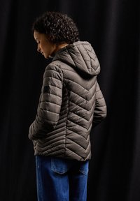Veste matelassée en gris foncé avec un motif en chevrons et une capuche. Comprend une fermeture éclair et des poignets élastiques, associée à un jean bleu.