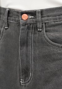 Κοντινή λήψη γκρι τζιν με λευκά ράμματα και πορτοκαλί κουμπί με ανάγλυφη επιγραφή "be" στη μέση.