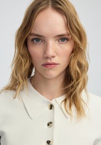 Camisa blanca de cuello con tejido texturizado, con grandes botones redondos oscuros. El modelo tiene el pelo ondulado a la altura de los hombros. Fondo neutro.