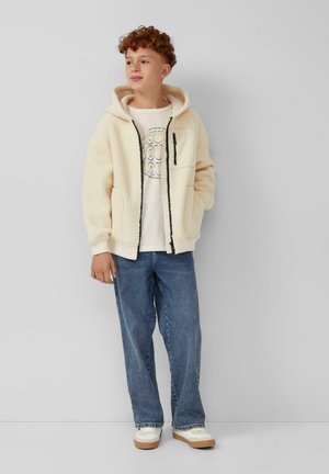 Fleece jack in crèmekleur met een ritssluiting, twee voorzakken en een capuchon, gecombineerd met een graphic wit T-shirt en blauwe jeans.