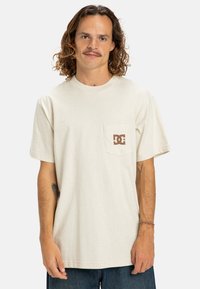 Krémové tričko s krátkým rukávem vyrobené z bavlny, s kapsičkou na levé straně hrudi a hnědým logem DC. Uvolněný střih a jemná textura.