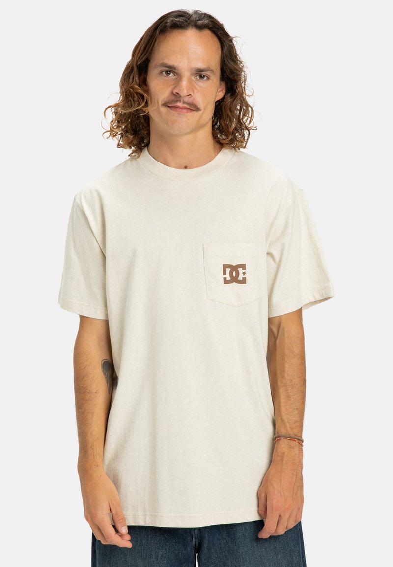 Krémové tričko s krátkým rukávem vyrobené z bavlny, s kapsičkou na levé straně hrudi a hnědým logem DC. Uvolněný střih a jemná textura.