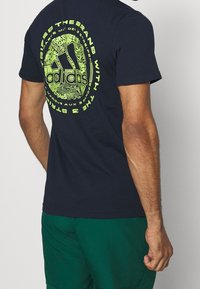 Tmavě modré bavlněné tričko s velkým neonově zeleným kruhovým potiskem s textem "adidas" a složitými vzory na zadní straně. Krátké rukávy.