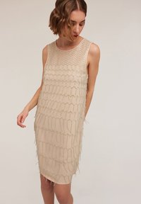 Nacktes ärmelloses Kleid mit strukturiertem Design, verziert mit gewellten, bestickten Akzenten und einer leicht durchsichtigen Schicht, die eine figurbetonte Silhouette betont.