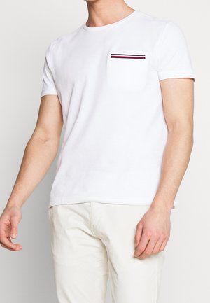 Uomo che indossa una maglietta bianca a maniche corte con una piccola tasca sul petto caratterizzata da strisce nere, rosse e bianche.