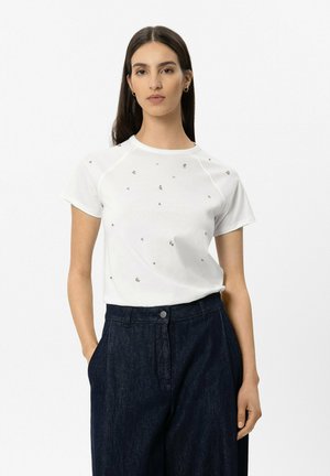 Woman with long dark hair wears white short-sleeve shirt with small crystal details and dark high-waist wide-leg pants, hands in pockets.