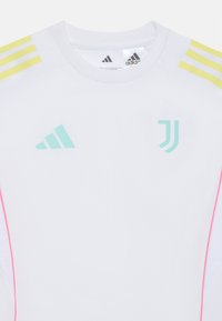 Camisa deportiva blanca con acentos en amarillo y turquesa, ribete rosa y logotipos. Presenta un cuello redondo y mangas cortas. Está hecha de un material ligero.