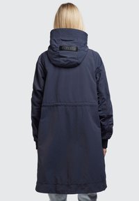 Granatowa parka z kapturem, z elastycznymi detalami, z naszywką logo na kołnierzu oraz średniej długości krojem. Gładki, lekki materiał.
