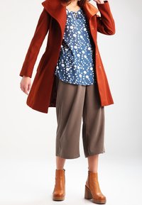 Cappotto color ruggine con un grande collo, camicia blu navy con pois bianchi e pantaloni grigi a lunghezza cropped. Stivaletti alla caviglia con tacco impilato marroni completano il look.