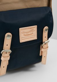 Granatowy i beżowy plecak wykonany z trwałej tkaniny, z charakterystyczną skórzaną łatą, metalowymi elementami i skórzanymi paskami z regulowanymi klamrami.