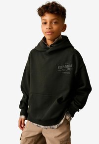 Čierna mikina s klokaním vreckom, na prednej strane s potlačou "SUPERDRY". Nosená cez kockovanú flanelovú košeľu, vyrobená z jemnej tkaniny.