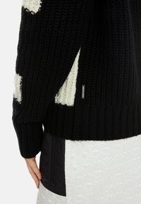 Schwarzer Strickpullover mit weißen strukturierten Patches, versehen mit einem gerippten Saum und einem Logo-Tag. Über einem strukturierten, hellen Kleidungsstück getragen.