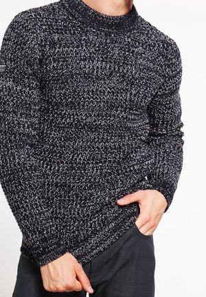 Homme portant un pull en maille à motif noir et blanc, une main dans une poche sombre et l’autre ajustant l’ourlet du pull.