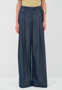 Pantaloni in denim scuro a gamba ampia con pieghe, doppie tasche frontali e cuciture a contrasto. Design a vita alta. Texture liscia.