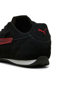 Schwarzer Wildledersneaker mit rotem Puma-Logo an der Ferse und roten Streifendetails, weiße Sohle, von hinten auf weißem Hintergrund gezeigt.