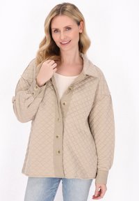 Beige gesteppte Jacke mit einem Kragen, großen Knöpfen und einem lässigen Schnitt, kombiniert mit einem leichten Oberteil und Jeans.