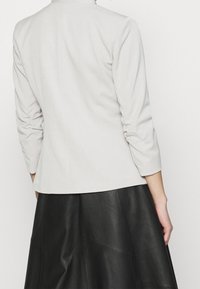 Jasnoszara, dopasowana marynarka z trójkątnymi rękawami i marszczeniami na ramionach, zestawiona z czarną skórzaną spódnicą.