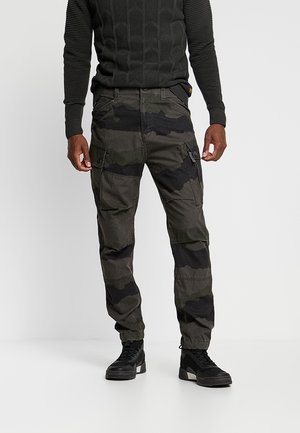 Mand iført mørke camouflage cargobukser, sort tekstureret sweater og sorte høje sneakers stående mod en ensfarvet baggrund.