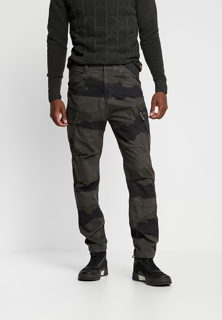 Homme portant un pantalon cargo camouflage foncé, un pull noir texturé et des baskets montantes noires, debout devant un fond uni.