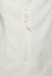 Biała kurtka z polaru z suwakiem z przodu. Suwak wyposażony jest w mały uchwyt wykonany z dopasowanego materiału. Miękki, teksturowany materiał.