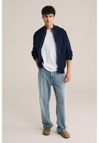 Mörkblå stickad cardigan med knappstängning, bärs över en vit t-shirt, kombinerad med ljusblå jeans och bruna slip-on skor.