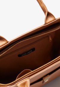Bolso de mano de cuero sintético marrón claro con herrajes en tono dorado, cierre con cremallera y bolsillo interior, que cuenta con una etiqueta de logo en su interior. Textura suave.