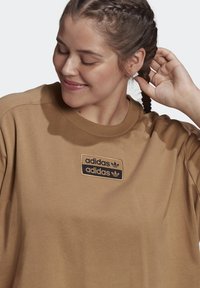 Bruine katoenen T-shirt met korte mouwen, voorzien van een zwart Adidas logo patch op de borst. Zachte textuur; ontspannen pasvorm ontwerp.