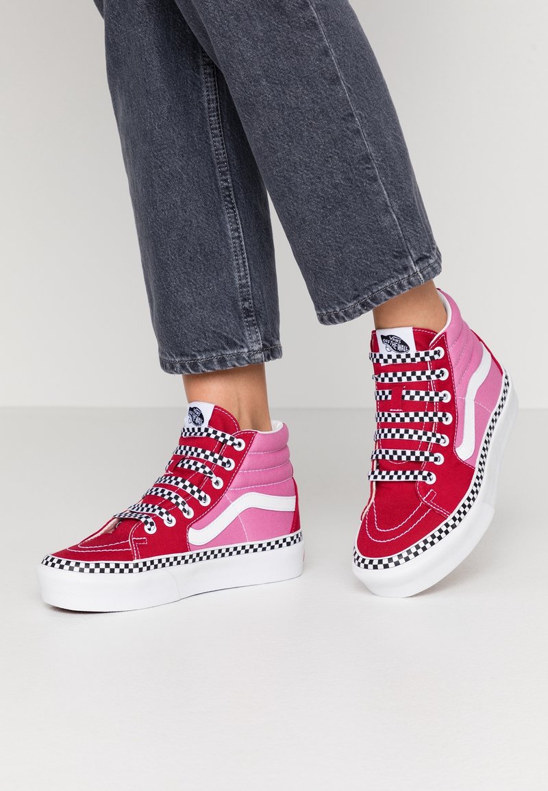 Personne portant des baskets montantes Vans rouges et roses avec des lacets et semelles à damier, associées à un jean denim gris foncé raccourci.