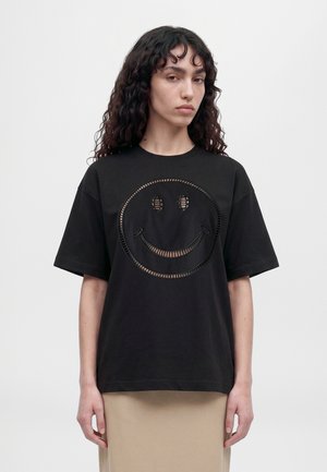 Un mannequin aux longs cheveux foncés et bouclés porte un T-shirt noir orné d’un grand smiley décoré, assorti à une jupe beige.