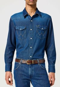 Dunkelblaue Jeanshemd mit spitzem Yoke, zwei Brusttaschen und weißen Knöpfen. Kombiniert mit einem braunen strukturierten Gürtel und blauen Jeans.