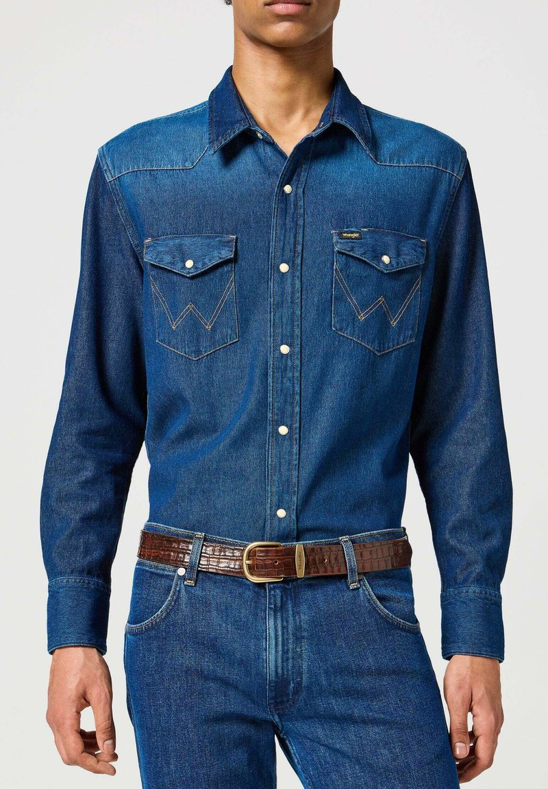 Dunkelblaue Jeanshemd mit spitzem Yoke, zwei Brusttaschen und weißen Knöpfen. Kombiniert mit einem braunen strukturierten Gürtel und blauen Jeans.