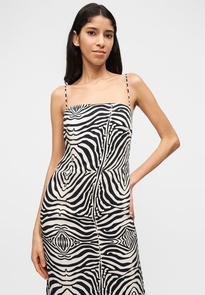 Γυναίκα με μακριά σκουρόχρωμα μαλλιά που φοράει ένα αμάνικο φόρεμα με μαύρο και άσπρο αφηρημένο μοτίβο και λεπτές τιράντες, ποζάροντας με το ένα χέρι στη μέση της.