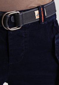 Detailní záběr na námořnické kalhoty se šedým tkaným pásem s dvojitým D-kroužkem a látkovou etiketou s brandingovým textem.