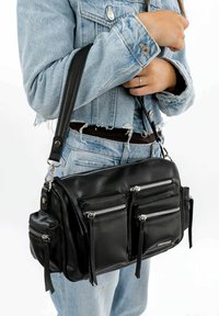 Black leather crossbody bag featuring multiple zippered pockets, smooth texture, and adjustable strap, carried alongside a denim jacket.