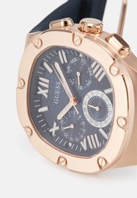 Montre en or rose avec un cadran noir texturé, des index horaires en chiffres romains et des sous-cadrans pour le jour et l'heure au format 24 heures. Bracelet en caoutchouc.
