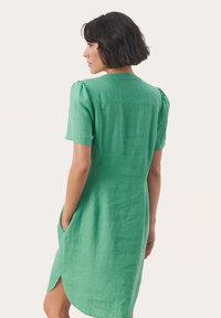 Grön linnklänning med korta puffärmar, bakstycke, sidofickor och en något rundad nederkant. Mjuk textur, avslappnad design.