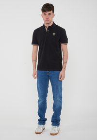 Polo nero con colletto accentato in oro, logo a scudo sul petto, abbinato a jeans blu e sneakers bianche con dettagli blu chiaro.