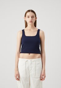Crop top bleu marine avec des bretelles larges et un décolleté carré. Tissu doux avec une coupe ajustée. Porté avec un pantalon cargo blanc ample.