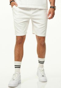 Weiße Shorts mit einem strukturierten Rauten-Muster, seitlichen Taschen und einer lockeren Passform. Kombiniert mit weißen Sneakers und gestreiften Knöchel-Socken.